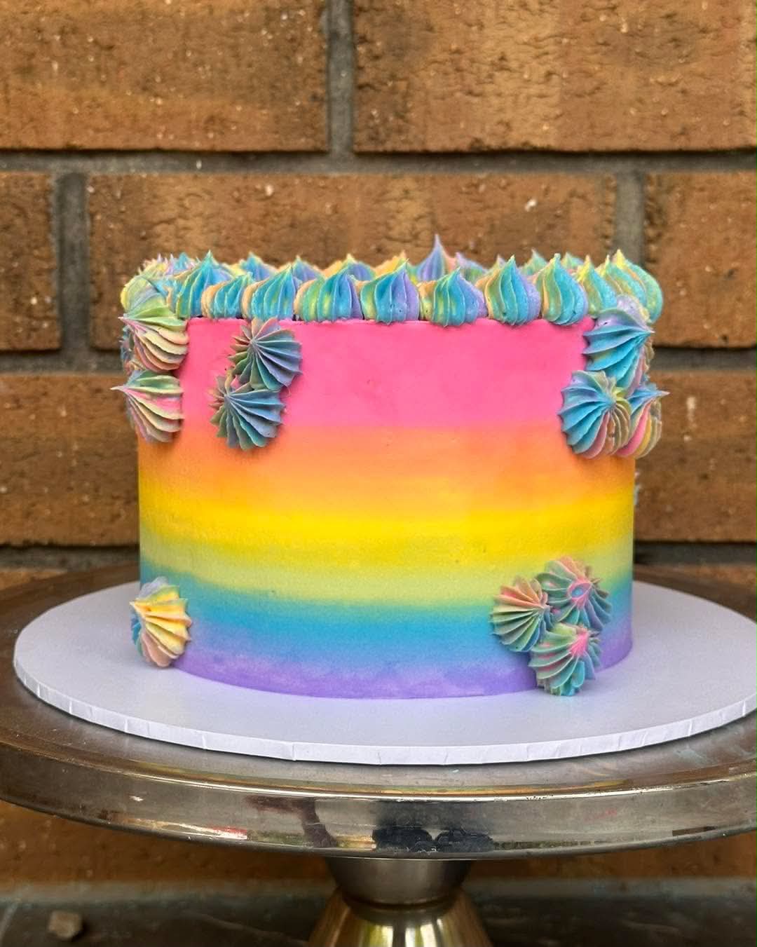 Rainbow ombre birthday cake with pink, orange, yellow, teal and purple buttercream, rainbow rosette border on top and multicolour pinwheel rosettes on sides